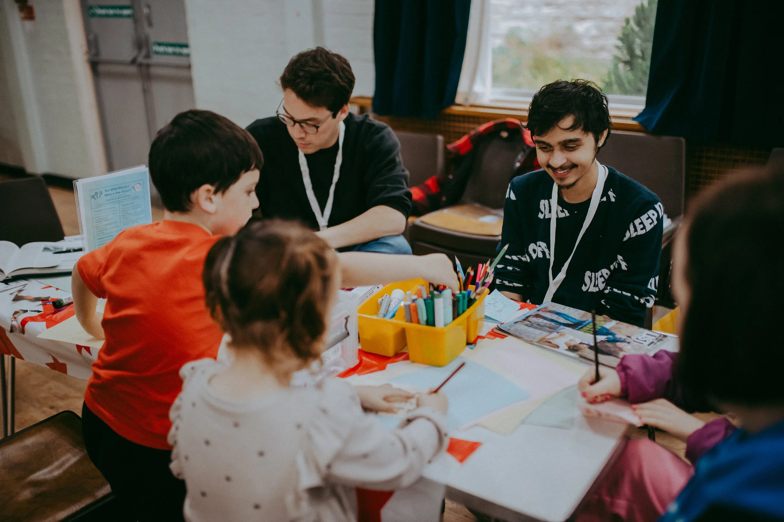 A group of children and teenagers engaged in arts and crafts activities around a table covered with art supplies, with a window and blue curtains in the background.