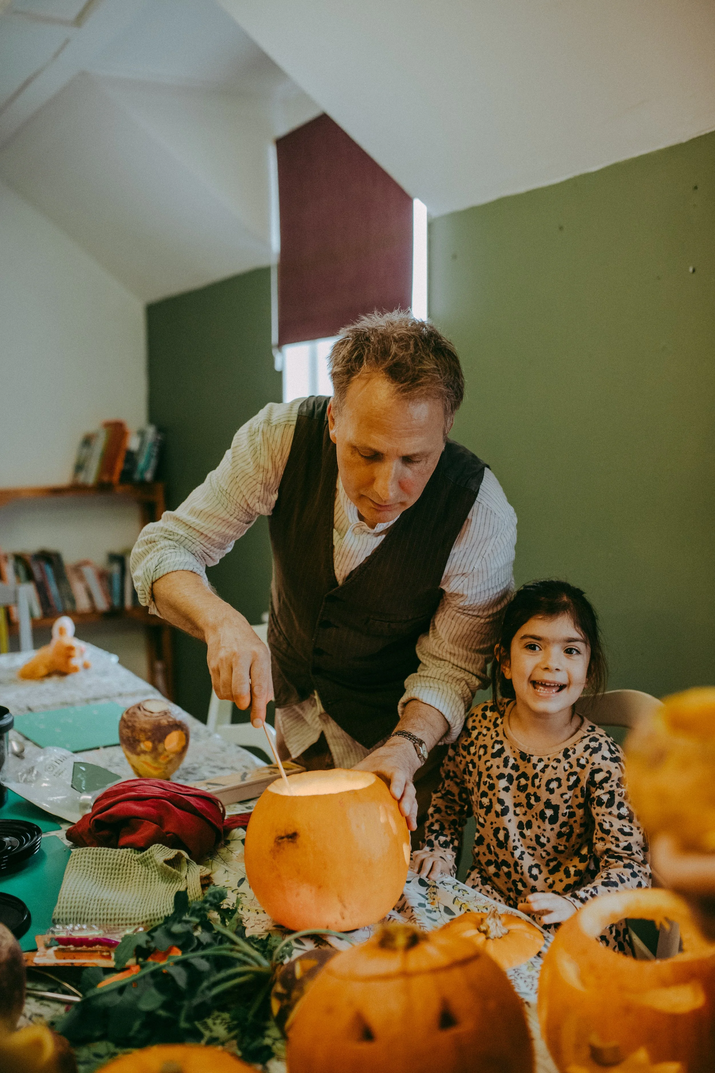 A man and young girl are carving a pumpkin at a table filled with various pumpkin decorations and carvings. The girl is smiling and wearing a leopard print shirt. The man is focused on carving the pumpkin.