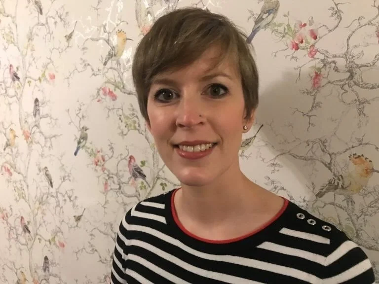 A fair skinned woman with short brown hair and a nose piercing smiling at the camera, wearing a black and white striped top with red trim, standing in front of a wallpaper with bird and tree branch illustrations.