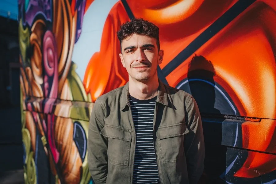 A fair skinned white man with dark, curly hair standing in front of a colorful graffiti mural on a wall, wearing a green jacket over a striped shirt, with sunlight casting shadows.