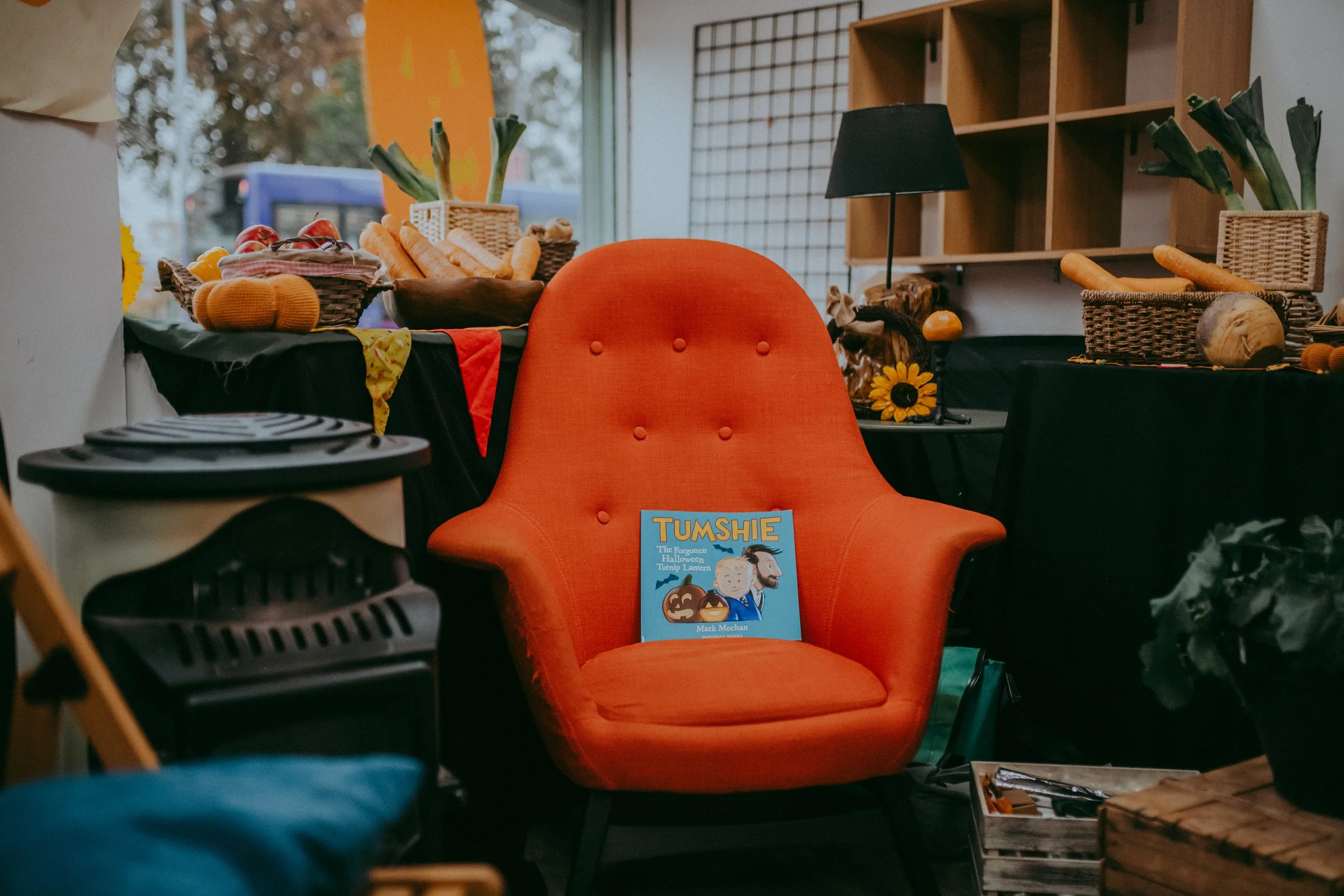 An orange cushioned chair with a book titled 'TUMSHIE' on it, placed in a room with wooden shelves and wicker baskets filled with vegetables like carrots and leeks, and other decorative items.