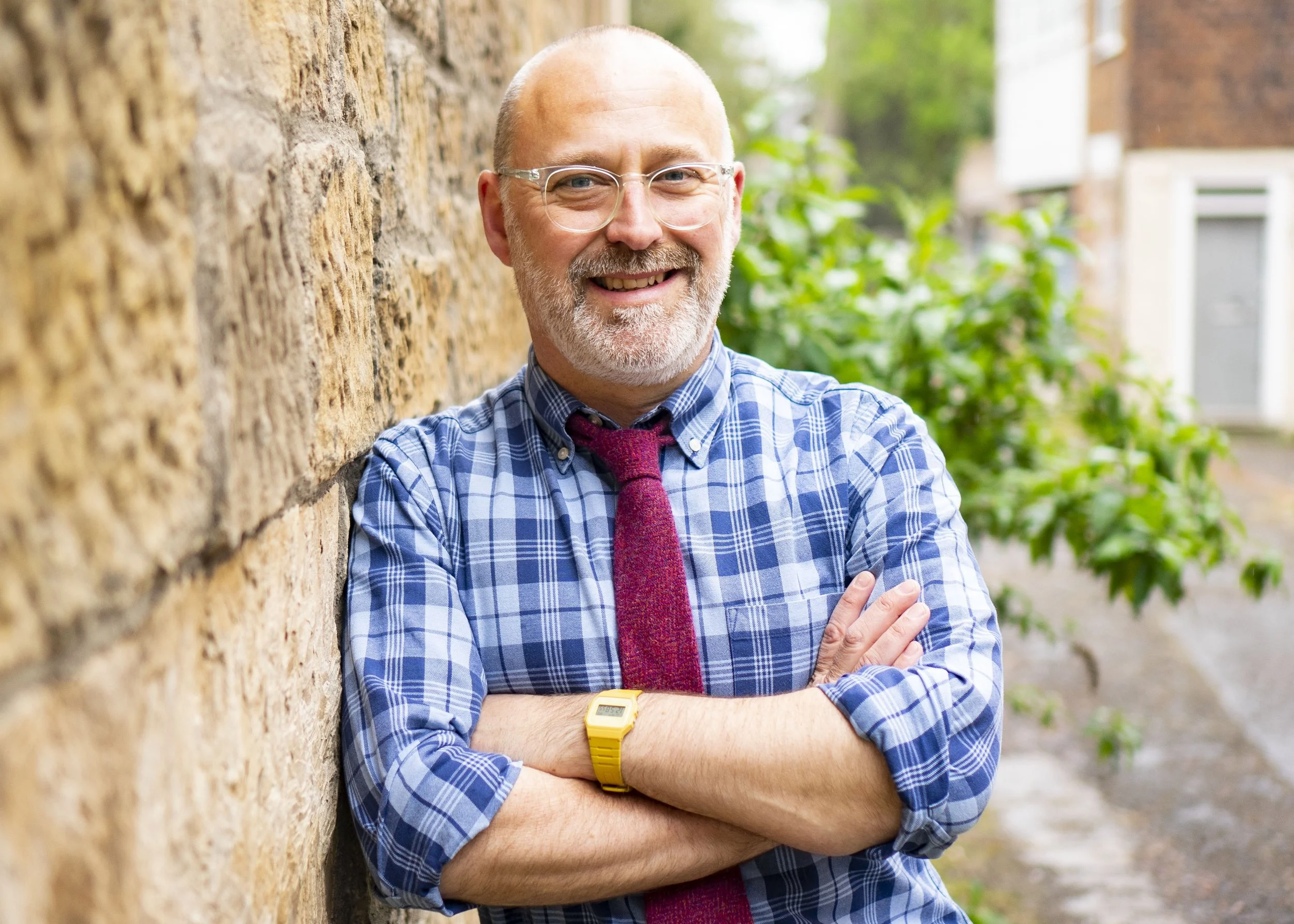 A smiling middle-aged man with glasses, a gray beard, wearing a blue plaid shirt, red tie, and yellow watch, standing outdoors beside a brick wall with green foliage in the background.