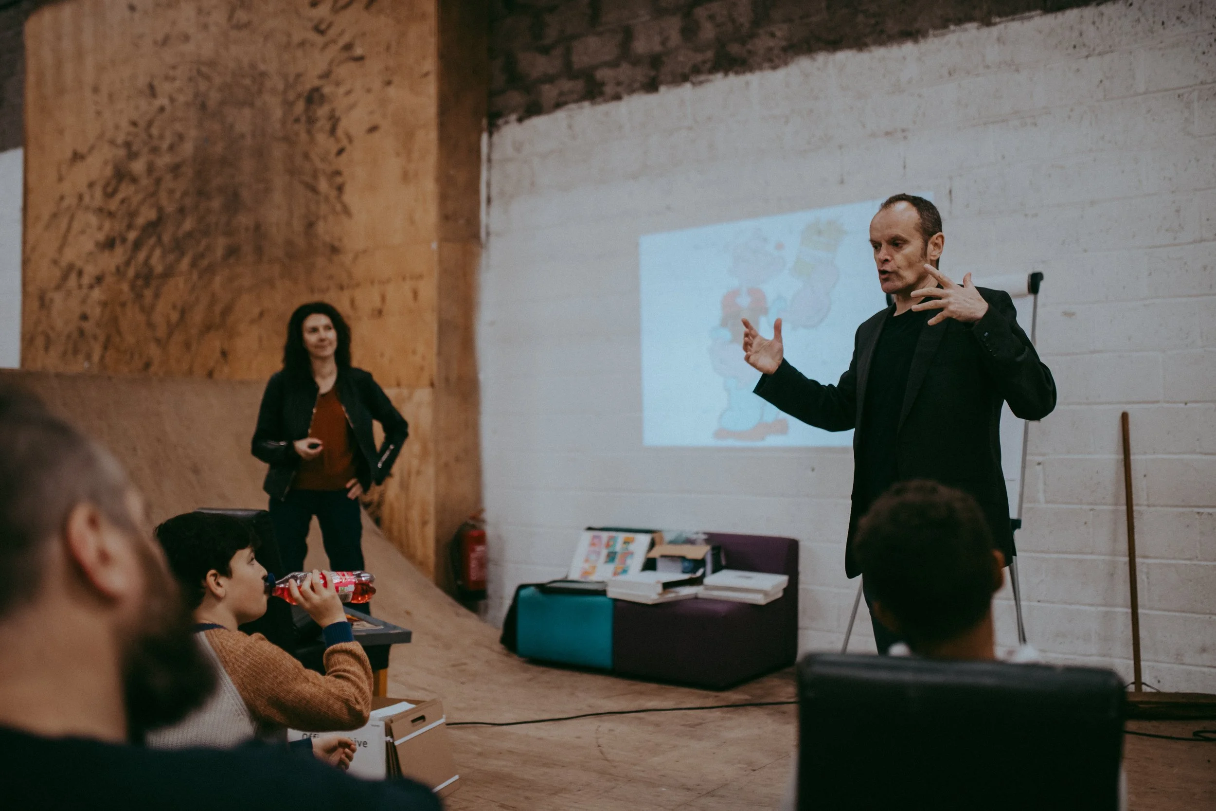 A man is giving a presentation indoors with a projected image on the wall behind him, while a woman stands nearby with her hands on her hips. Several children are seated, watching the presentation, with one child drinking from a bottle.