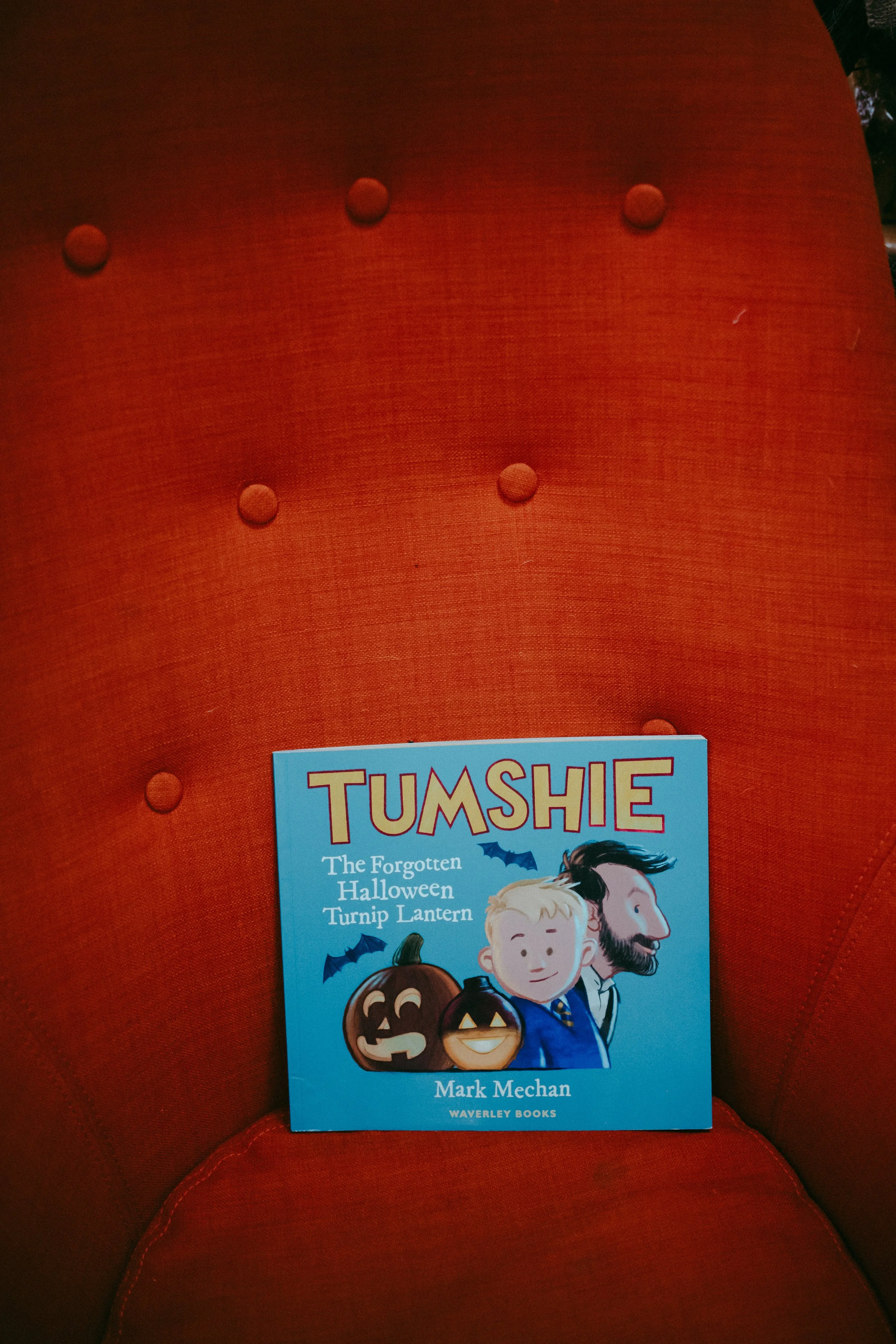 A blue children's book titled "TUMSHIE: The Forgotten Halloween Turnip Lantern" is displayed on a red upholstered chair, with part of the chair's curved backrest visible in the background.