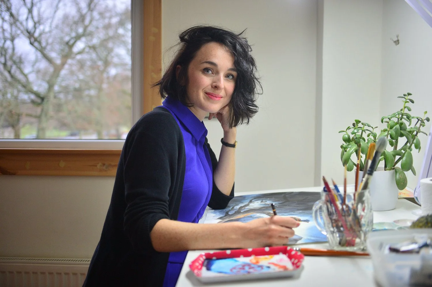 Fair skinned Woman with dark, curly hair sitting at a table, painting on canvas, smiling, with a potted plant and art supplies nearby, looking at the camera.