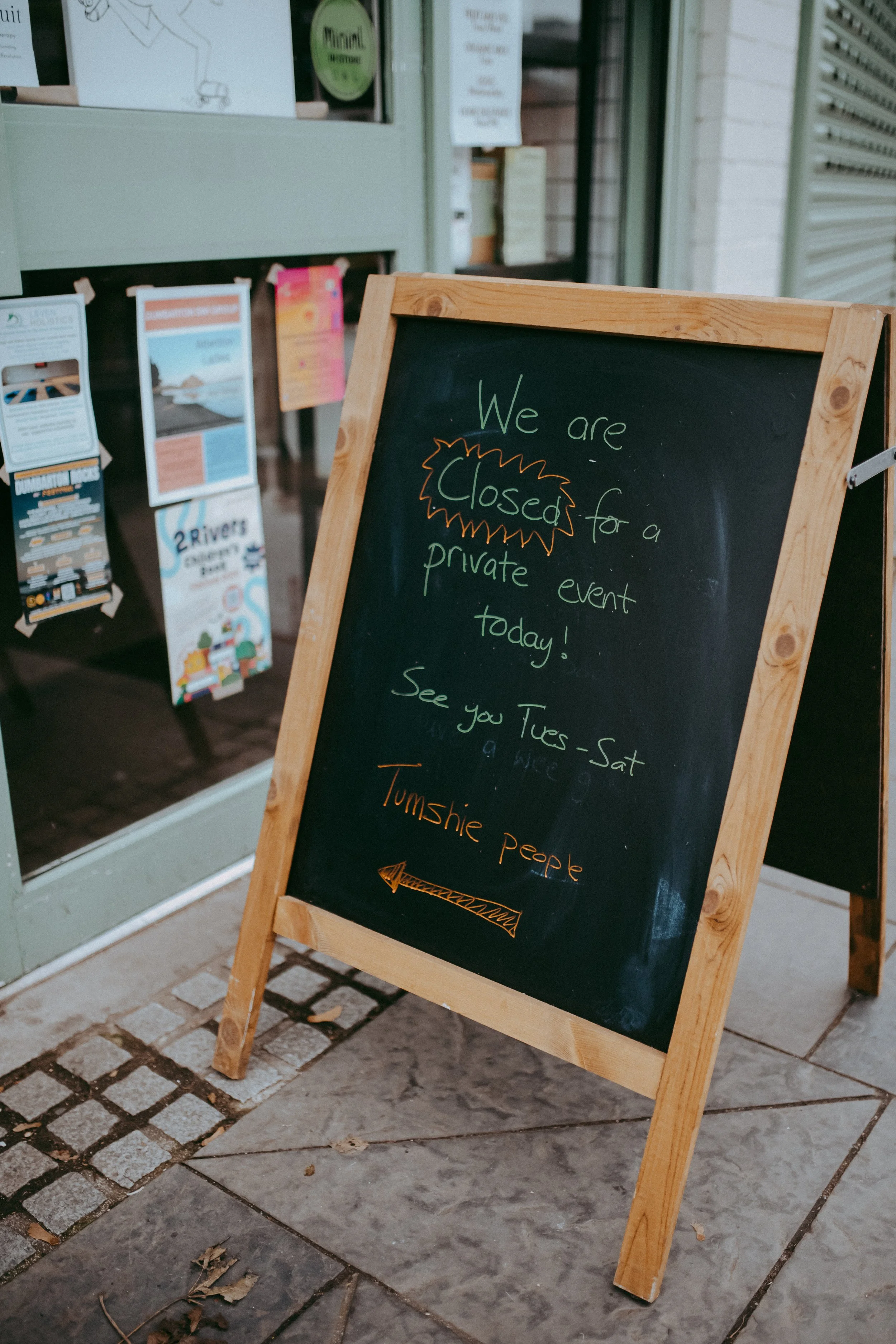 A black chalkboard sign with a wooden frame placed outside a store, displaying a message that reads: "We are closed for a private event today!" The sign mentions seeing customers on Tuesday through Saturday. In the background, there are posters and n