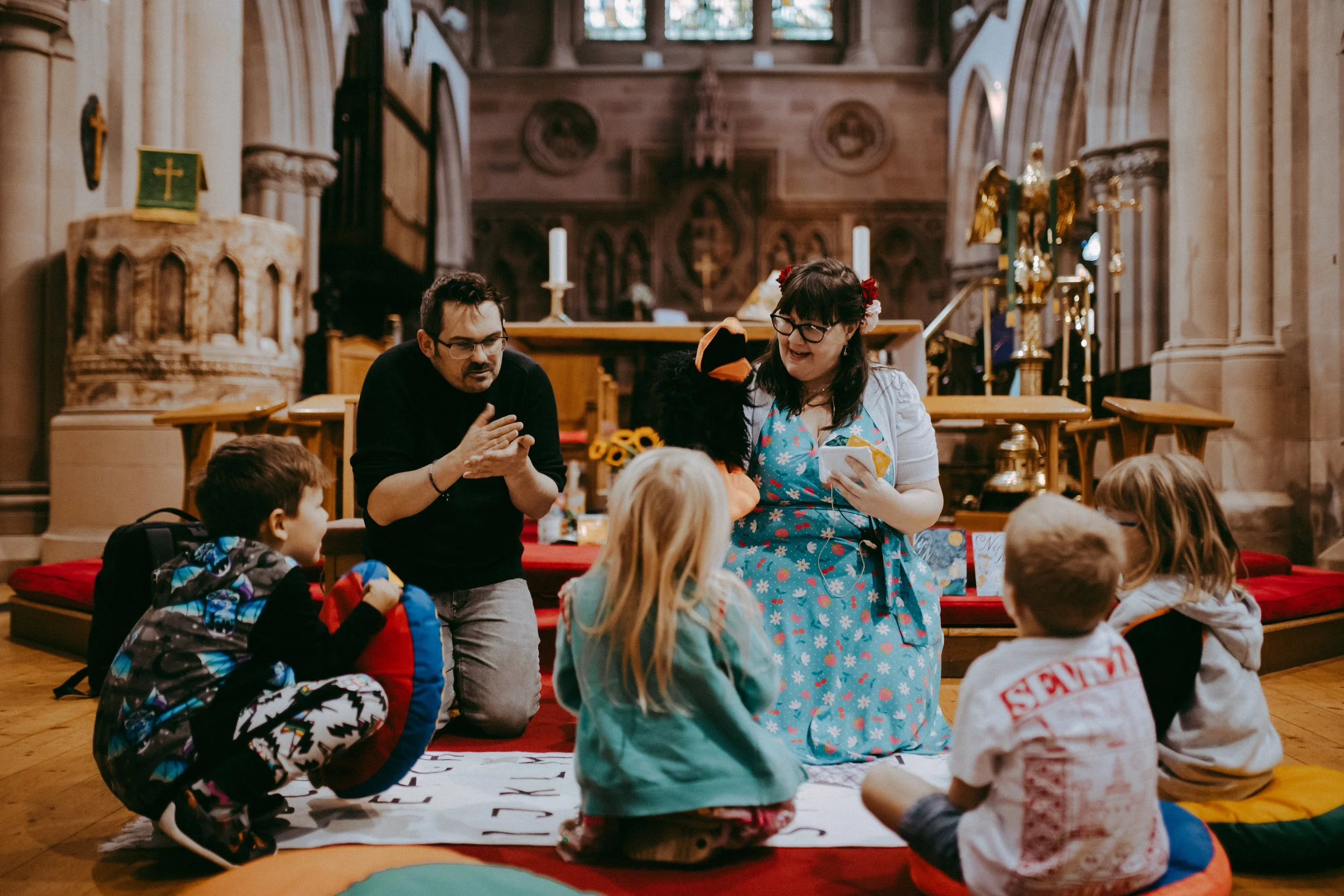 Children sitting on the floor of a church, listening to a woman dressed in a colorful apron, who is reading or performing for them. A man is communicating in sign language, and the church interior has wooden walls& stained glass windows
