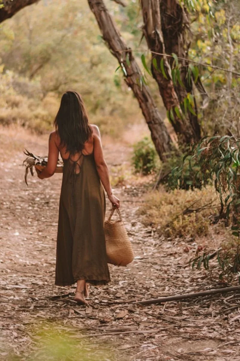 A woman with long dark hair wearing a sleeveless dress walking on a dirt trail through a wooded area, holding a basket in one hand and a bundle of leaves in the other.