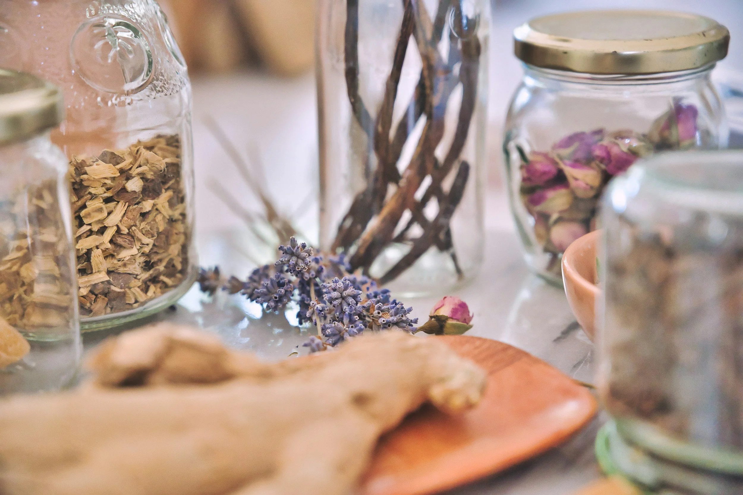 Close-up of glass jars filled with dried herbs, flowers, and botanicals on a table, with some dried flowers scattered around.