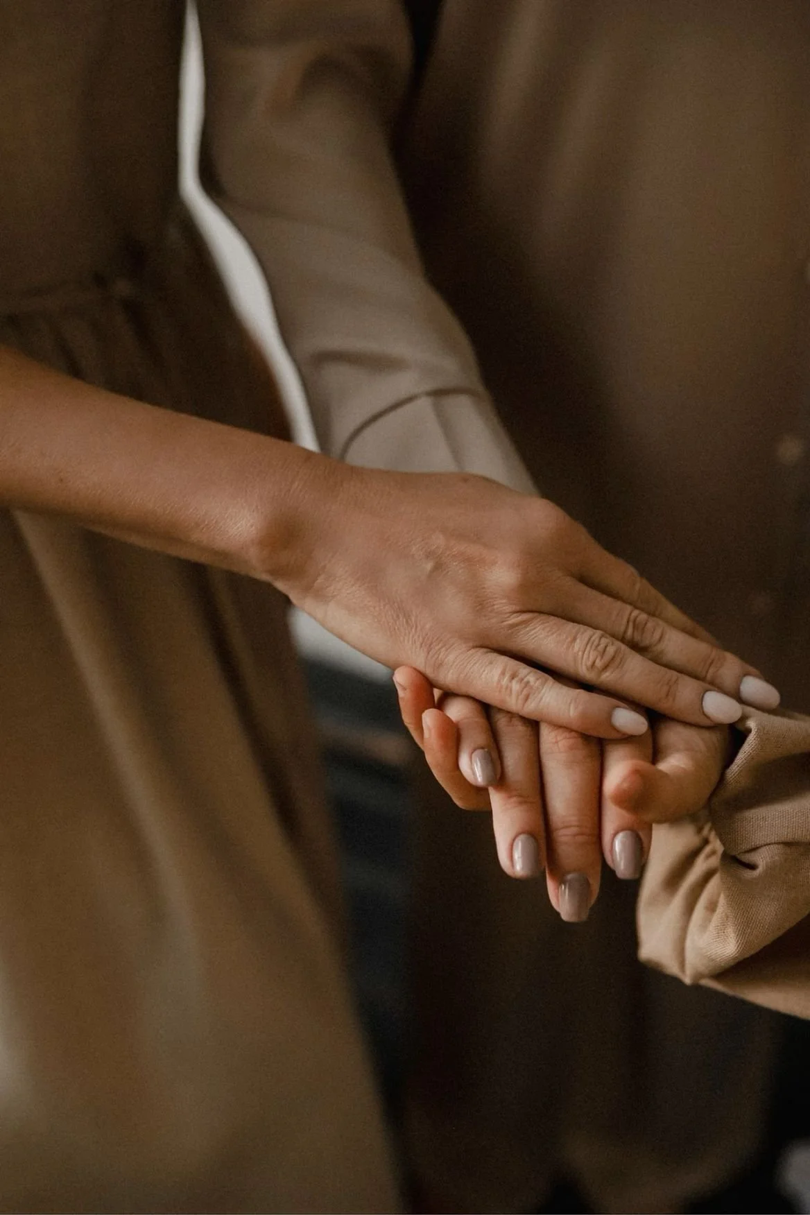 A close-up of two hands gently holding each other, with one person's hand resting on top of the other's, against a brown background.