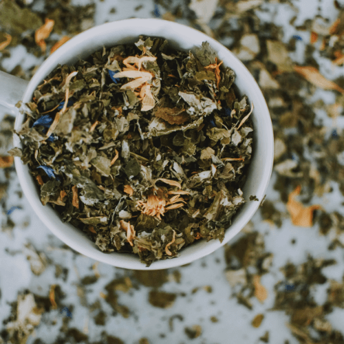 A white cup filled with loose leaf herbal tea surrounded by spilled tea leaves.