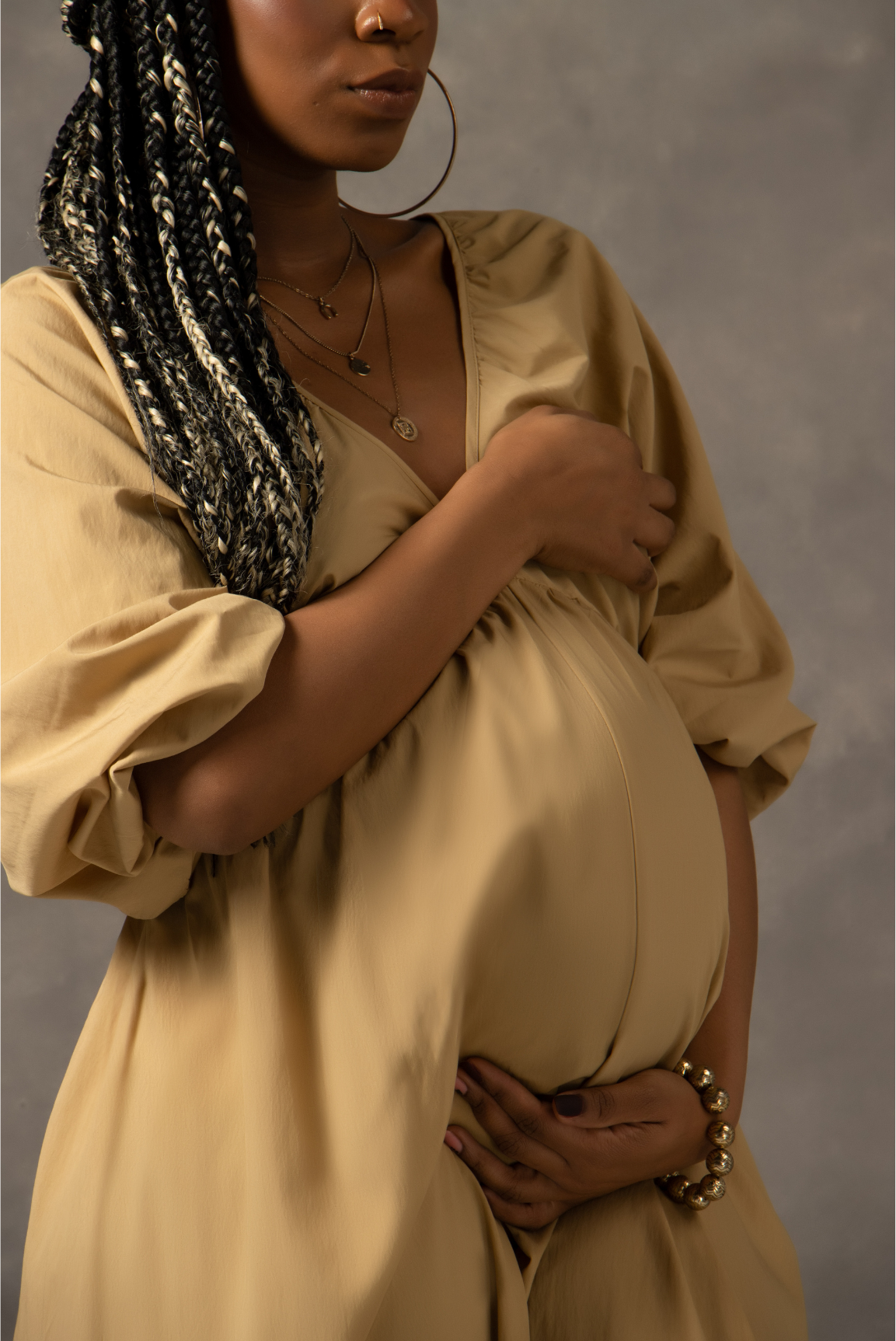 Pregnant woman in a beige dress with braided hair, wearing jewelry, covering her chest with one hand and holding her belly with the other, standing against a neutral background.