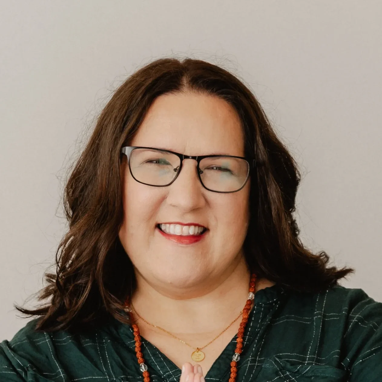 Portrait einer lächelnden Frau mit Brille, dunklem Haar und dunklem grünen Hemd vor hellem Hintergrund.