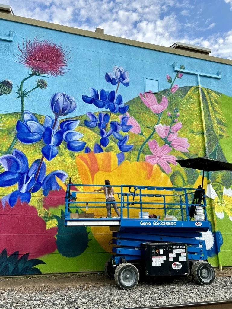 A mural depicting colorful flowers and green hills is being painted on a building's exterior wall. Two artists are working on the mural using a blue scissor lift, with one artist actively painting.