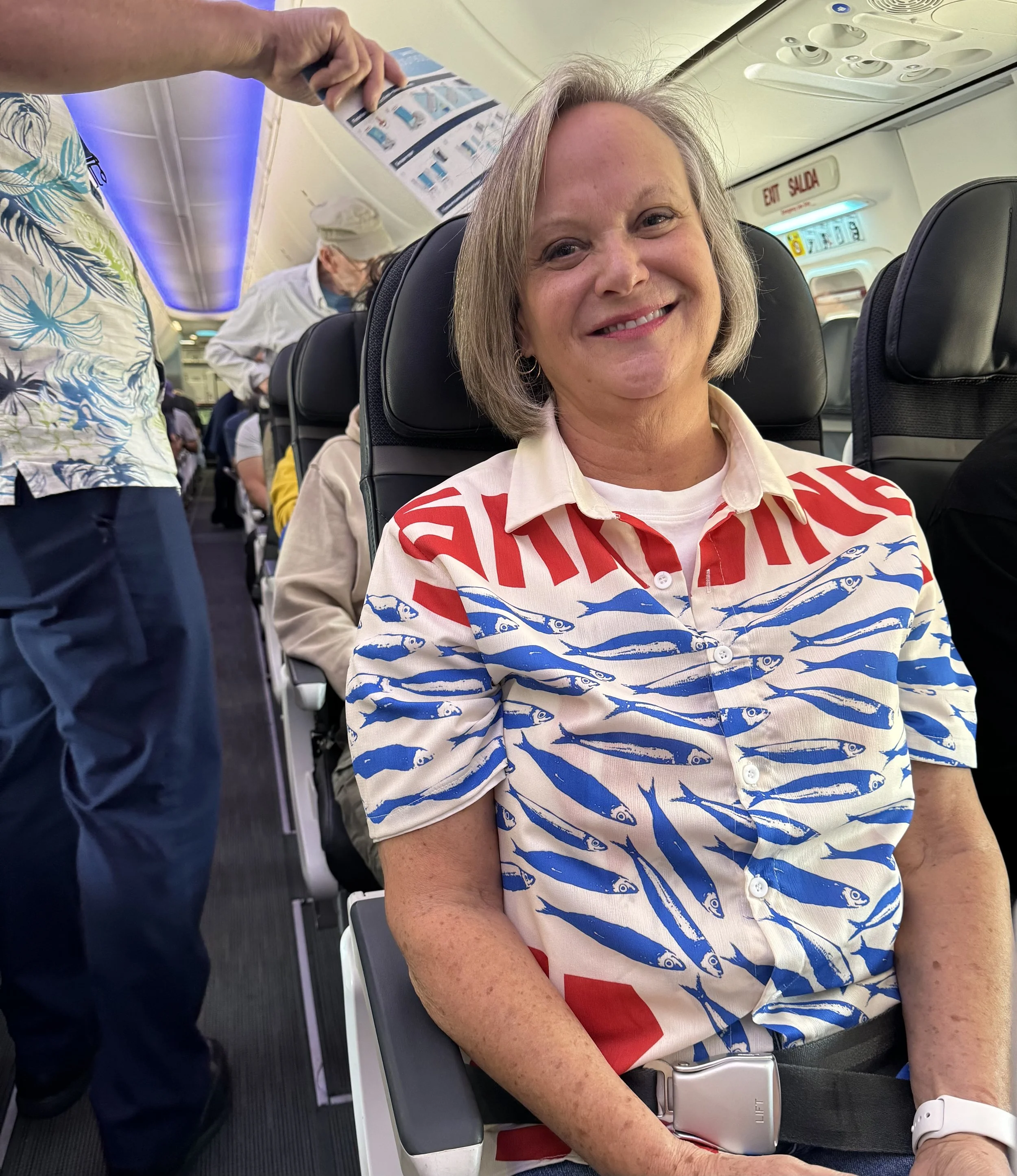 A woman sitting in an airplane seat, smiling, wearing a colorful shirt with a fish pattern and red and blue lettering, with other passengers and airplane interior visible in the background.