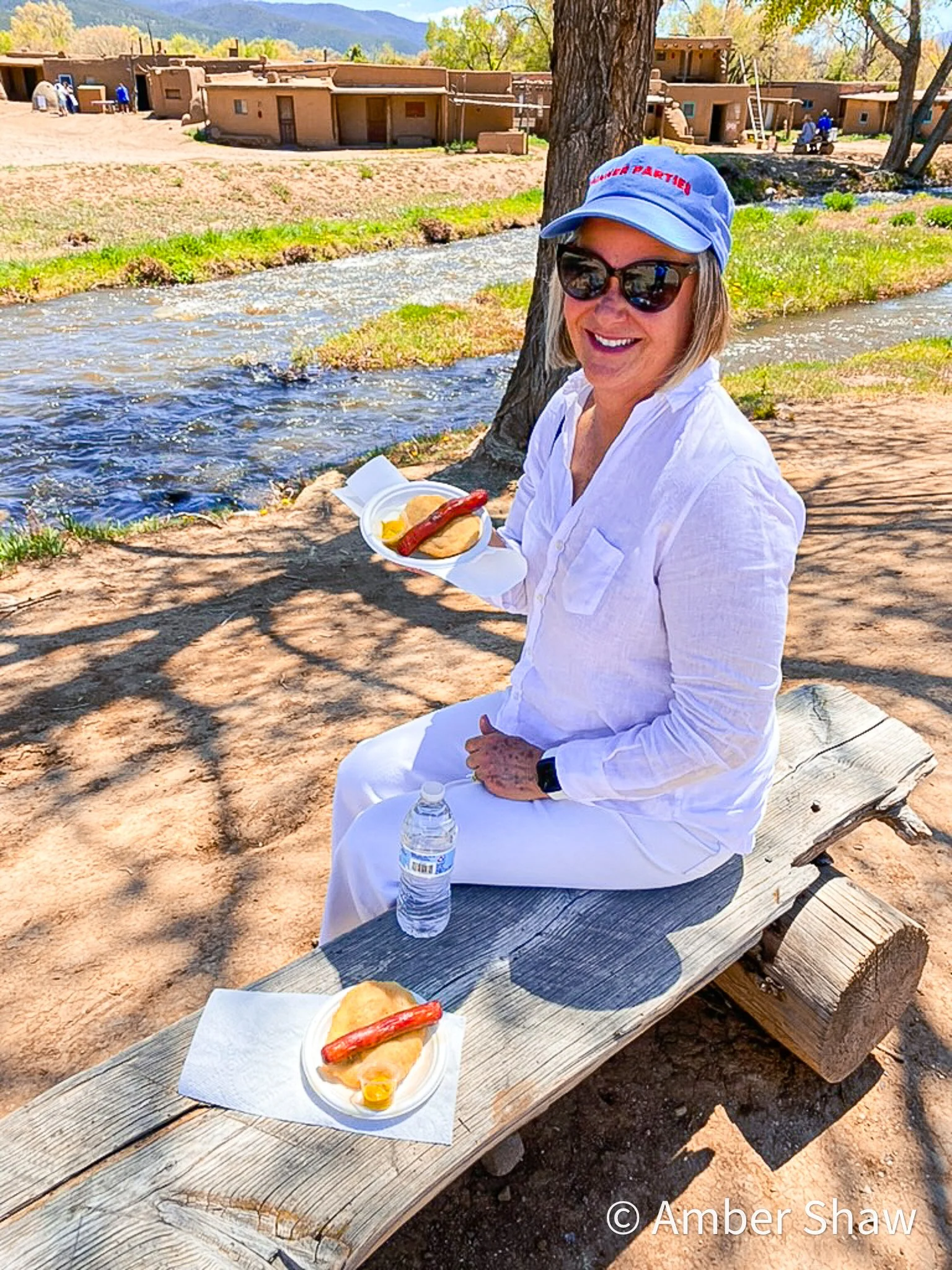 A woman sitting on a wooden bench outdoors near a river, holding a plate with a, and mustard. She is wearing a white outfit, sunglasses, and a blue hat, smiling at the camera. There is a water bottle on the bench in front of her, and a second plate of the same food on the bench. Trees and adobe-style buildings are visible in the background.