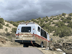 A bus parked on a dirt road in a rocky, hilly area with sparse trees under cloudy skies.