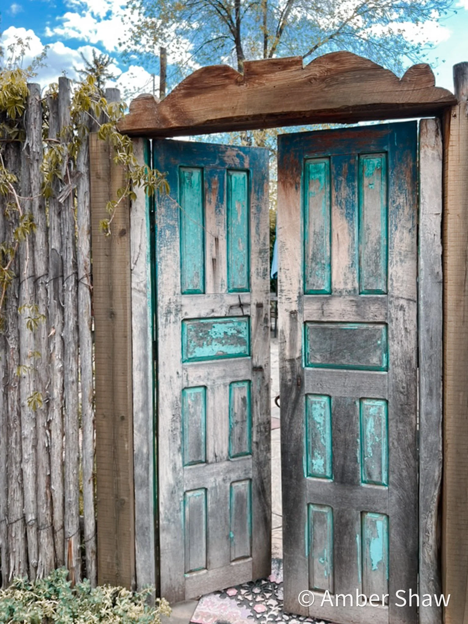 Open weathered wooden double doors with teal paint, set within a rustic fence and garden, under a blue sky with clouds.
