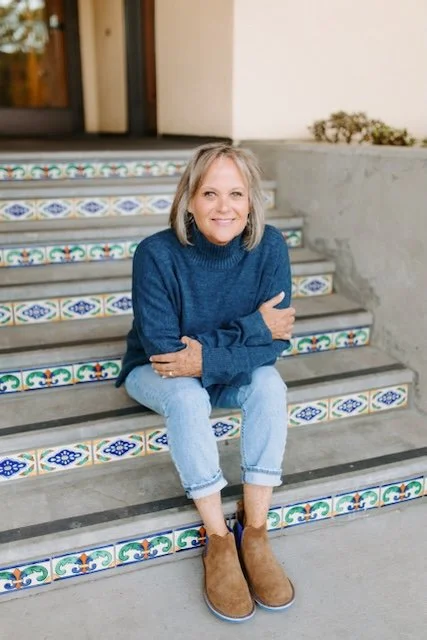 A woman with blonde hair sitting on outdoor decorative tiled stairs, wearing a blue sweater, light jeans, and brown boots, smiling at the camera.