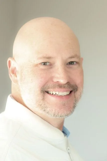 Close-up of a smiling bald man with light skin and a beard, wearing a white shirt.