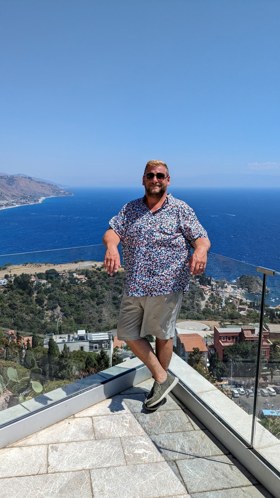 A man with beard and sunglasses wearing a patterned shirt and shorts standing on a glass platform overlooking the ocean and coastline with hills in the background.
