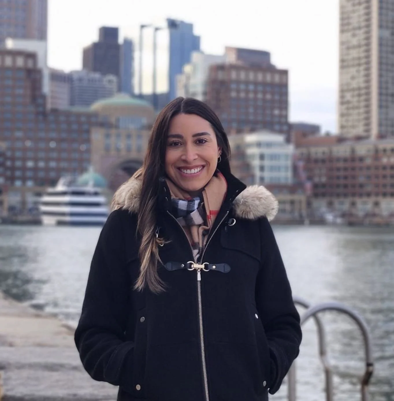 A woman smiling outdoors near a body of water with a city skyline in the background.