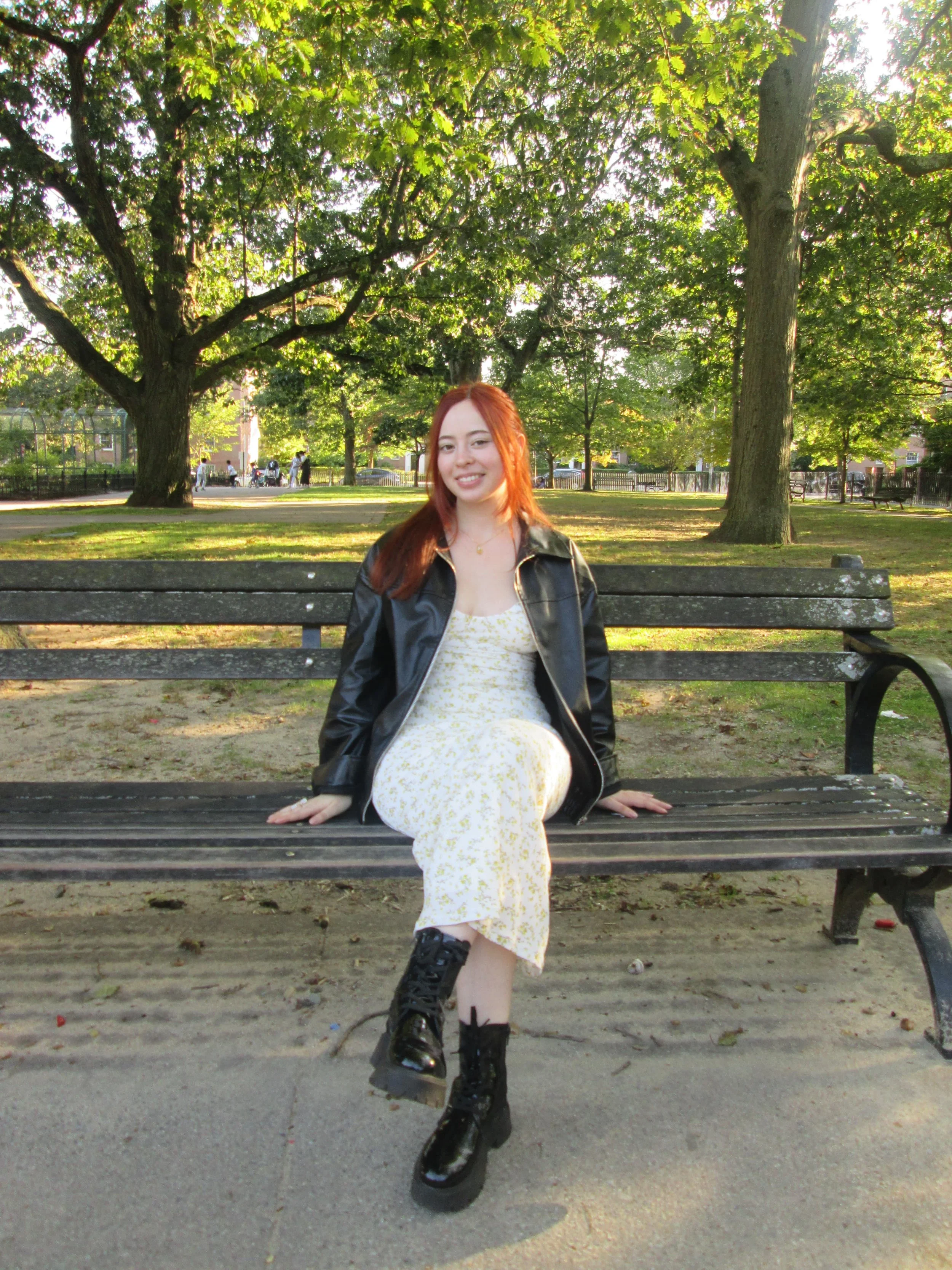 Young woman with red hair sitting on a park bench surrounded by green trees, wearing a yellow dress, black leather jacket, and combat boots.