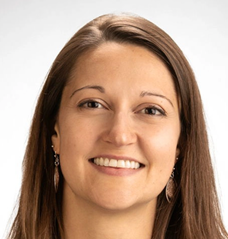 Close-up portrait of a smiling woman with long brown hair, wearing earrings, against a plain white background.
