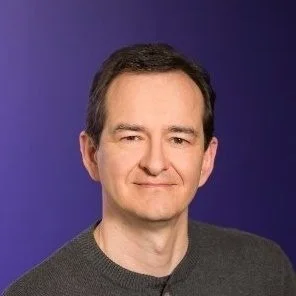 A man with short dark hair, wearing a gray sweater, smiling against a blue background.
