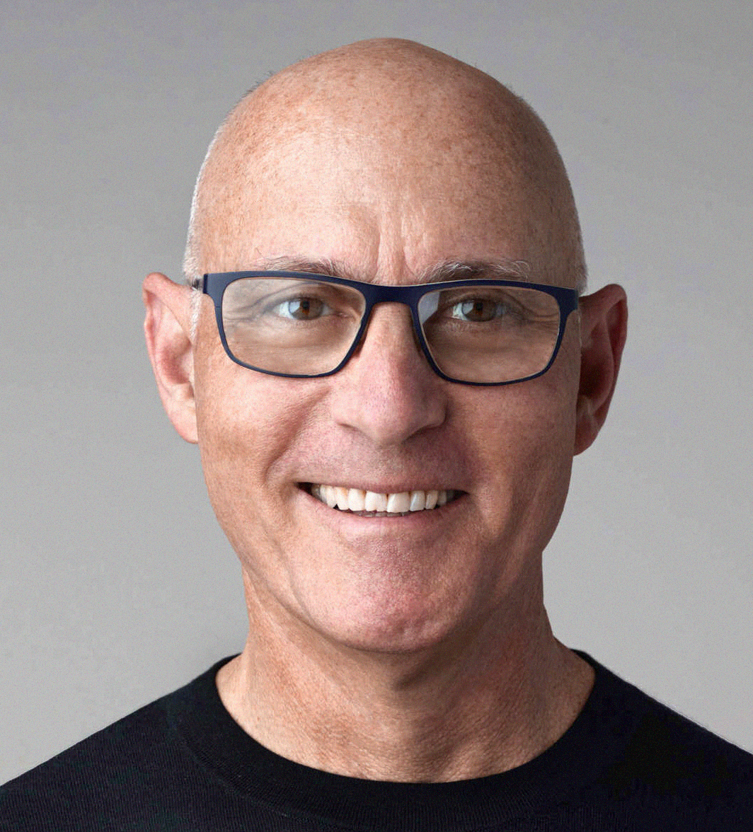 Close-up portrait of a smiling, bald man wearing dark glasses and a black shirt against a plain gray background.