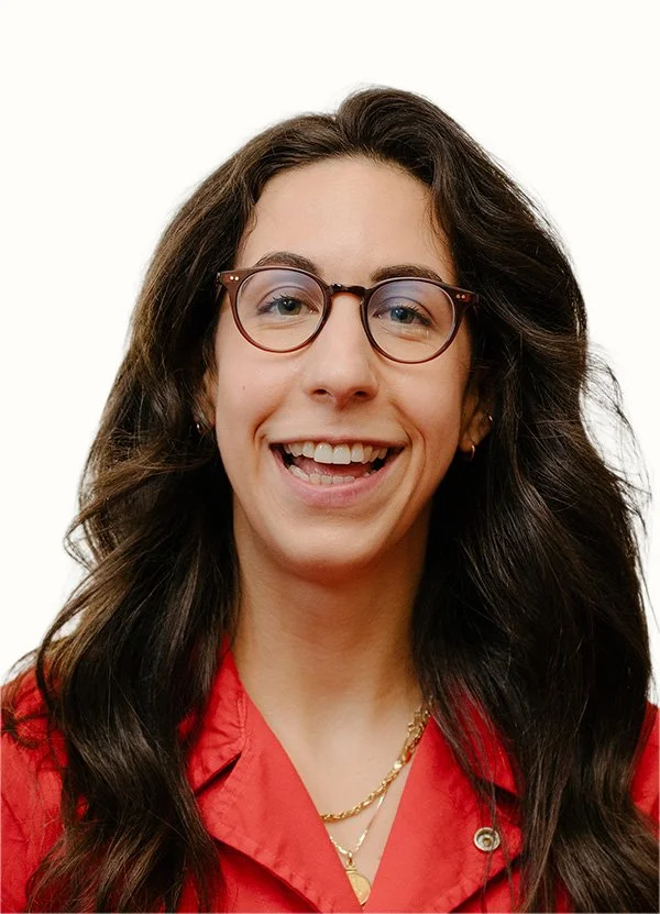 A young woman with long wavy brown hair, wearing glasses and a red blouse, smiling in front of a white background.