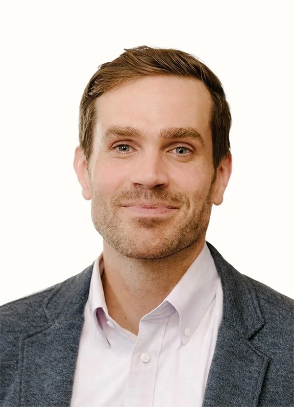 Headshot of a man with brown hair and a beard, wearing a white shirt and a dark grey blazer, smiling against a plain white background.