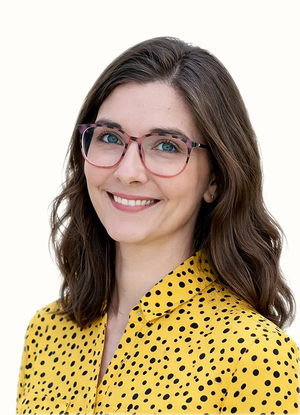 A woman wearing pink-rimmed glasses and a yellow polka dot blouse, smiling with shoulder-length wavy brown hair, against a plain white background.