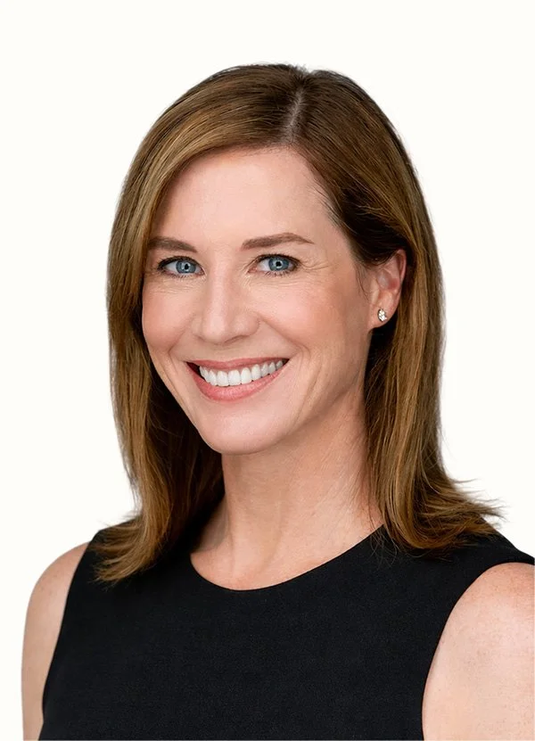 A woman with shoulder-length brown hair, blue eyes, and light skin, smiling and wearing a sleeveless black top against a white background.
