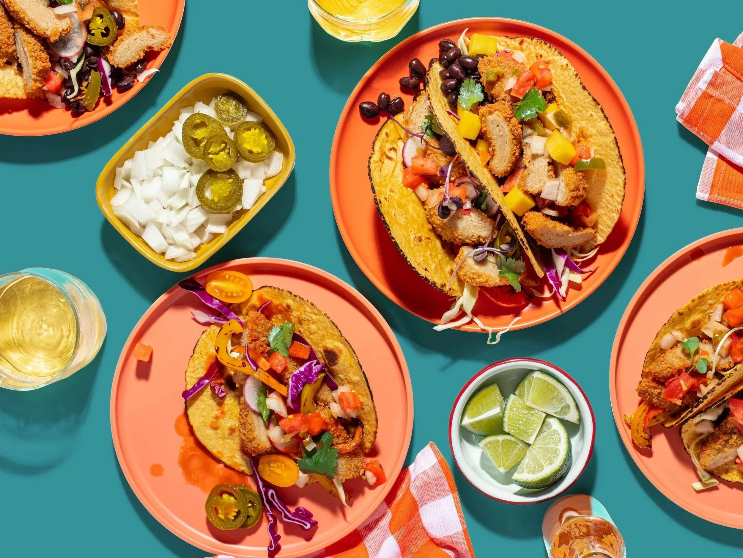 Several plates of prepared tacos filled with fried protein, topped with shredded vegetables and herbs, along with bowls of lime wedges, pickled onions, jalapeño slices, and drinks on a teal table with a checkered napkin.