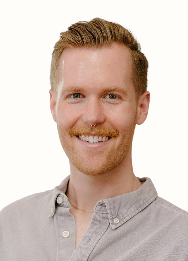 Portrait of a young man with ginger hair, a mustache, and a septum piercing, smiling while wearing a light gray button-up shirt.