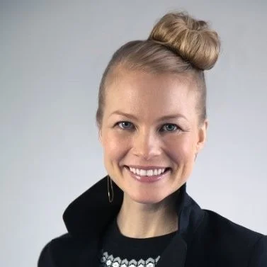A woman with blonde hair styled in a bun, smiling, wearing a black jacket against a plain background.