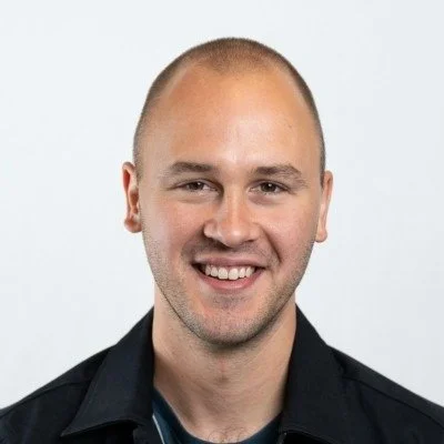A young man with a shaved head and a slight beard, smiling, wearing a dark shirt against a plain light background.