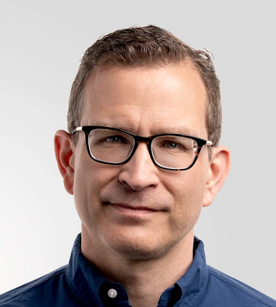 Portrait of a man with short brown hair, wearing black glasses and a navy blue shirt, looking at the camera against a plain gray background.