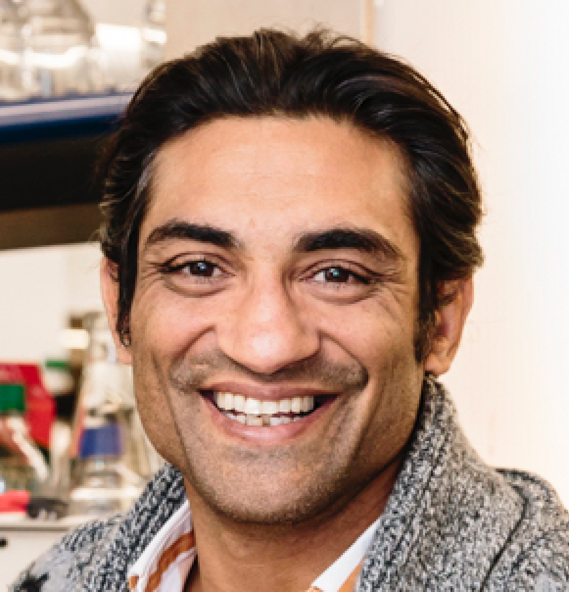 Close-up of a smiling man with dark hair, wearing a gray textured blazer, in an indoor setting with kitchen supplies in the background.