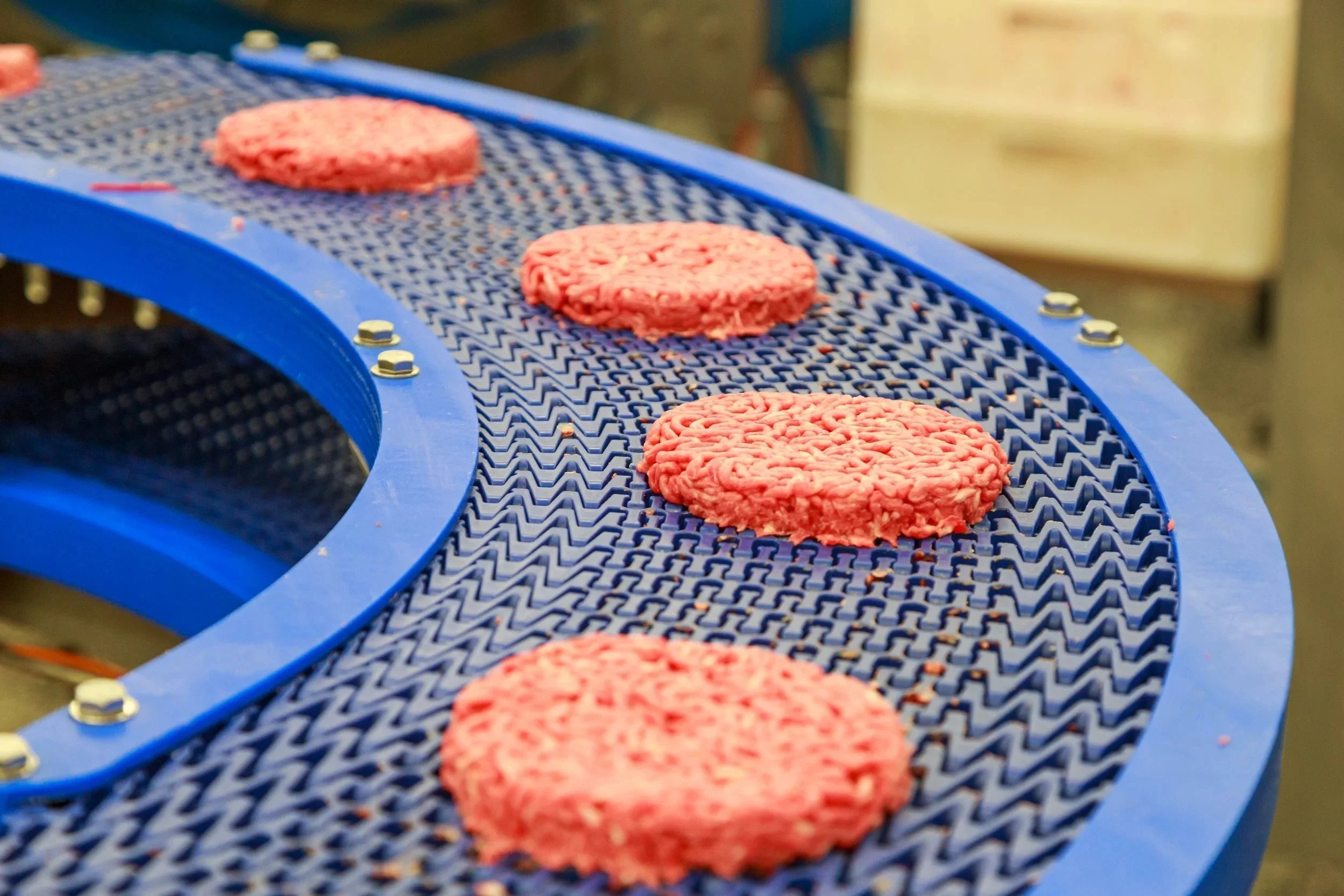 Pink alternative protein patties on a blue conveyor belt in a processing facility.