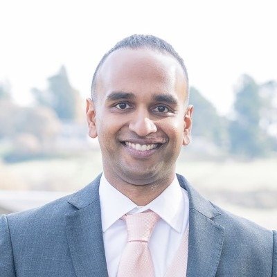 A smiling man in a suit and tie outdoors with blurred trees and sky in the background.