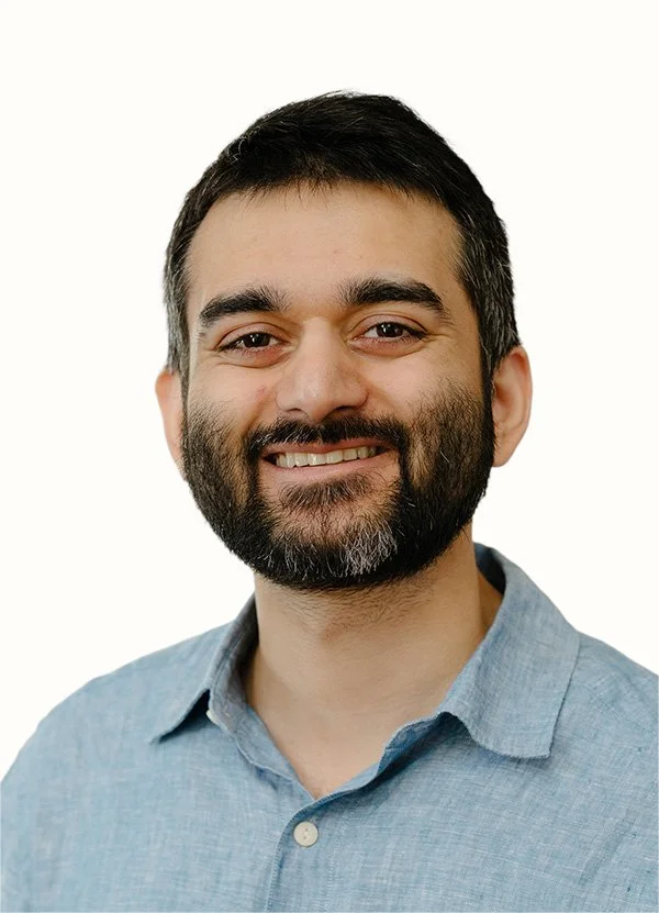 Close-up of a smiling man with dark hair, beard, wearing a light blue button-up shirt, against a plain white background.