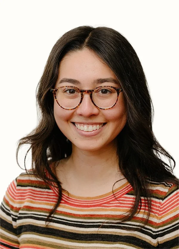 A young woman with shoulder-length dark hair, glasses, and a striped shirt, smiling against a white background.