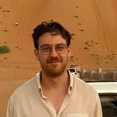 A man with curly dark hair, glasses, and a beard standing outdoors in front of a sandy, desert landscape with some vehicles in the background.