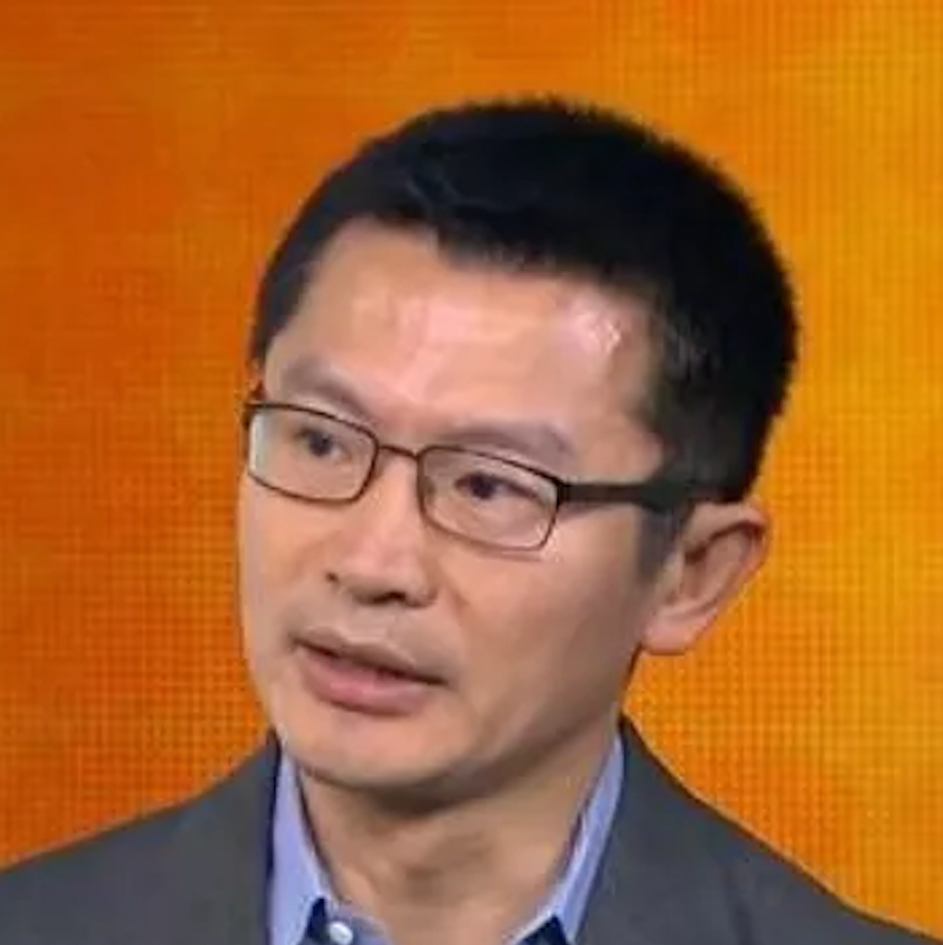 Man with black hair, glasses, and wearing a suit jacket, speaking during a televised interview against an orange background.