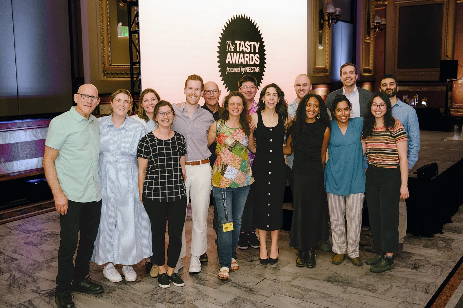Group of people posing together on stage at an awards event, with a large screen behind displaying 'The Tasty Awards powered by Nectar' logo.