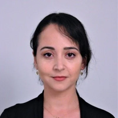 A woman with short dark hair, wearing a black blazer, smiling slightly against a plain light gray background.