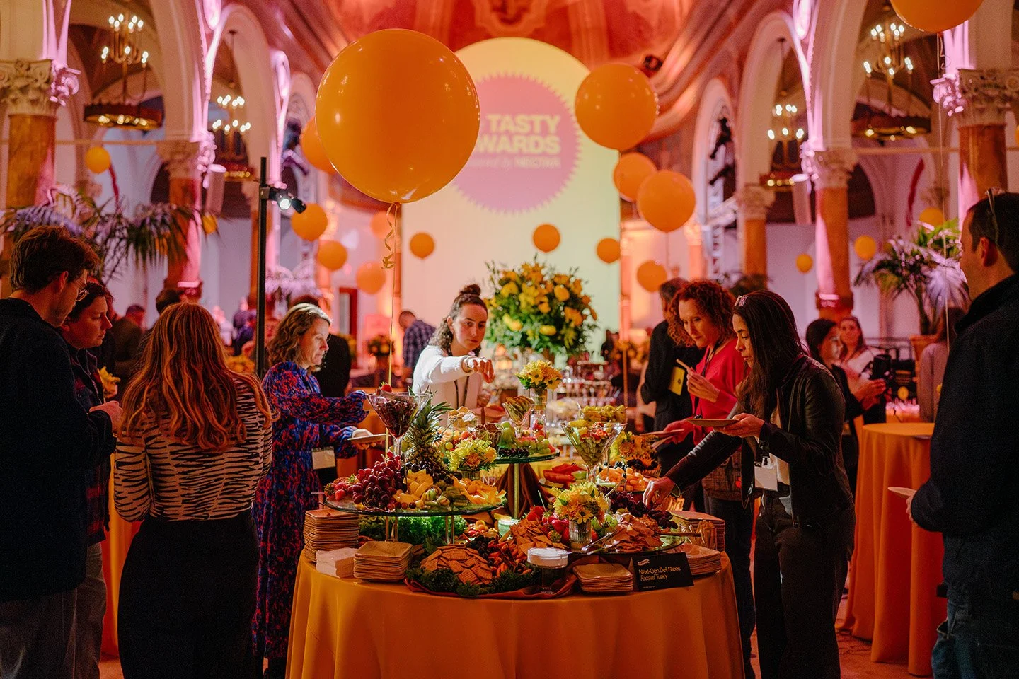 Guests at a buffet table with various food and snacks at an event with orange balloons and pink lighting.