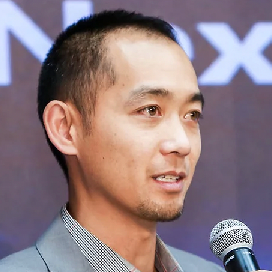 A young man with short black hair, wearing a gray blazer and checked shirt, speaking into a microphone at a public event.